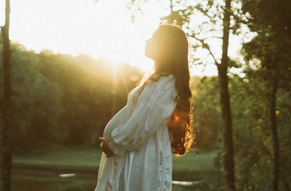 Pregnant woman in white dress enjoying a sunset in a serene forest setting.