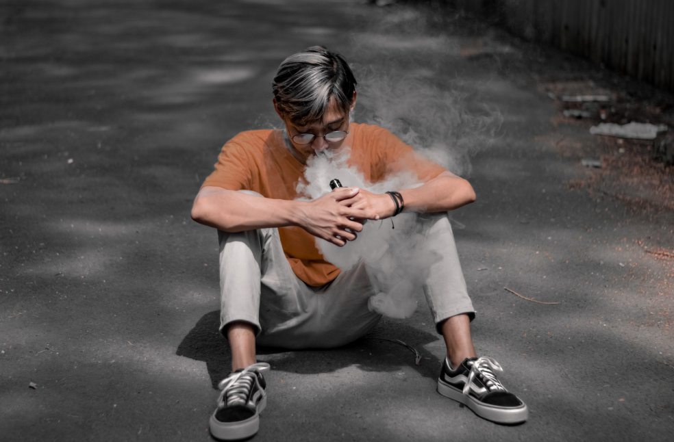 A young man wearing eyeglasses sits on an urban street while vaping