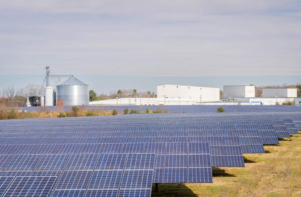 A large solar panel field with warehouses and silos in the background under a clear sky