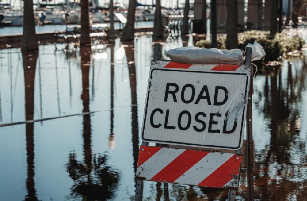 Road closed sign amidst flooded street, reflecting calm water and trees.