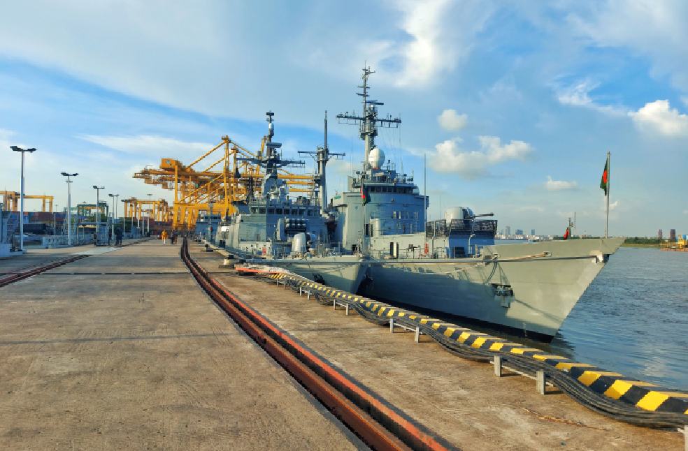 Navy vessels docked at a bustling city port with cranes and clear blue skies