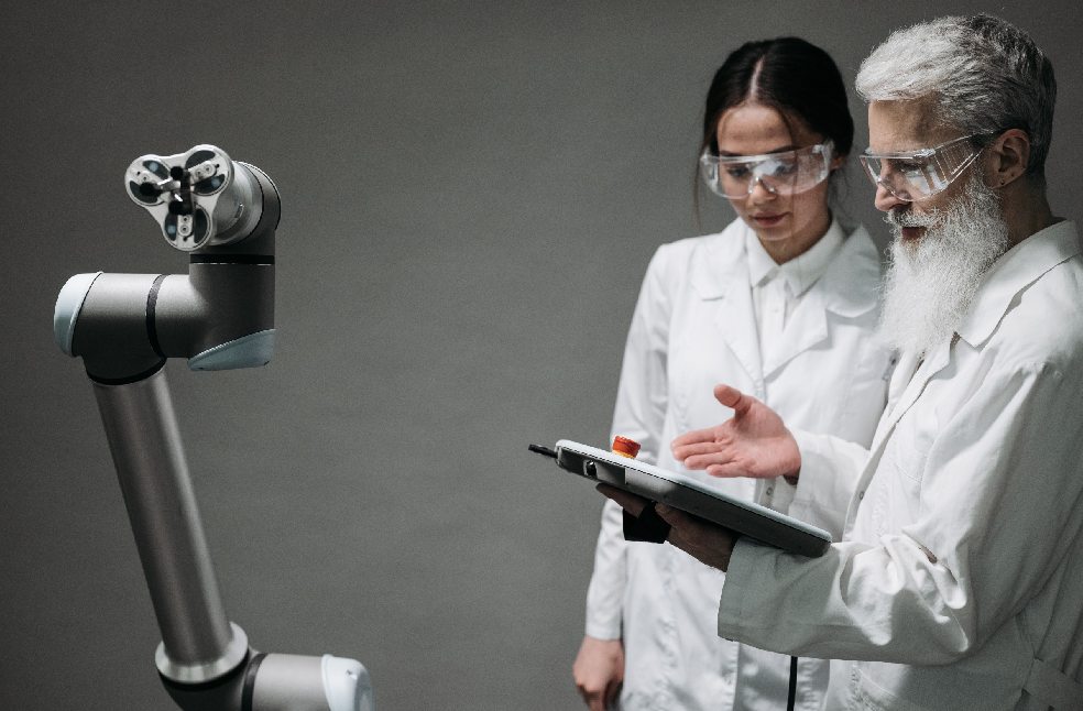 Researchers in lab coats and safety glasses engaging with a robotic arm in a lab setting