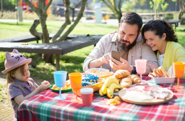Study links screen habits of parents to weaker family bonds Happy family having a picnic in a sunny park, enjoying snacks and using a smartphone together