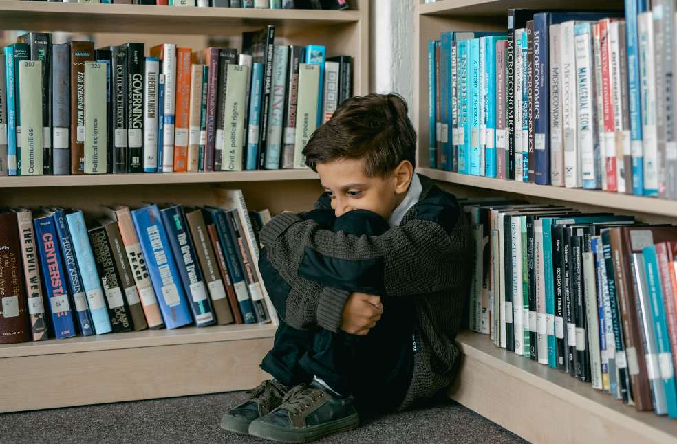 Young boy in library appears sad, sitting alone among bookshelves, suggesting themes of solitude and bullying.