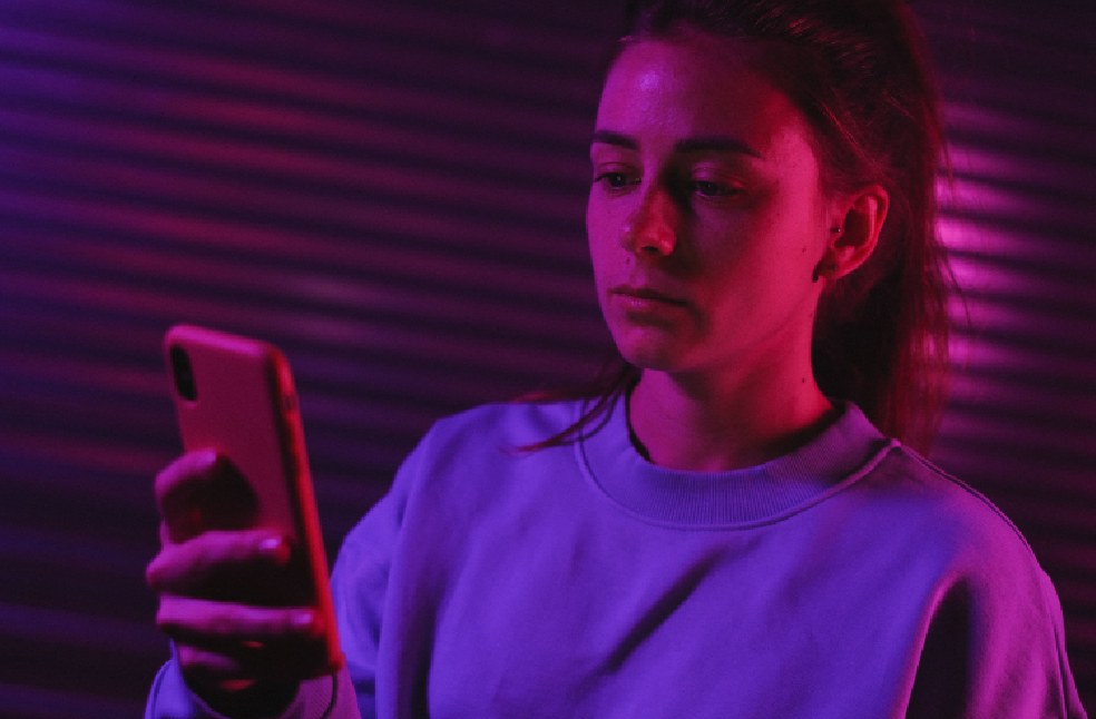 A young woman with a serious expression looks at her smartphone in a room illuminated by neon lights.