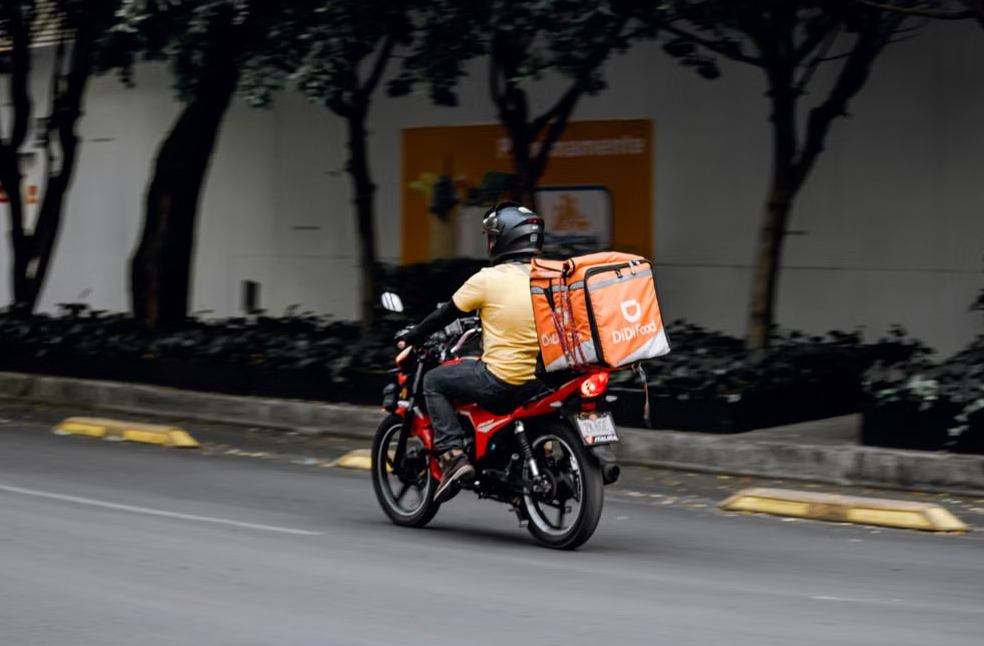 A delivery man riding a motorcycle with a thermal bag on a city street during the day