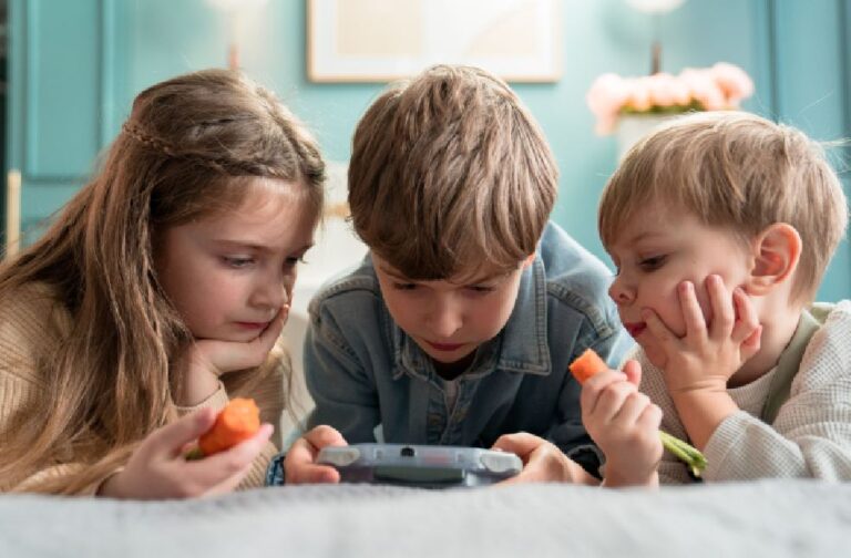 EU plans social media limits for children across Europe Three kids enjoying video games and snacking on carrots indoors, showcasing childhood togetherness