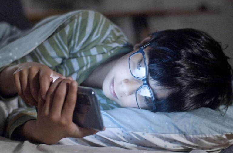 How blue light from screens disrupts sleep and eye health A child lying in bed using a smartphone at night, wearing glasses