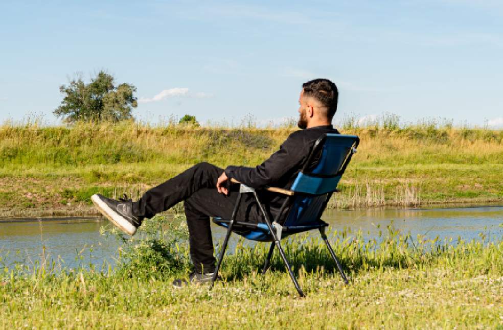 A man sits on a chair by a peaceful river enjoying a sunny day