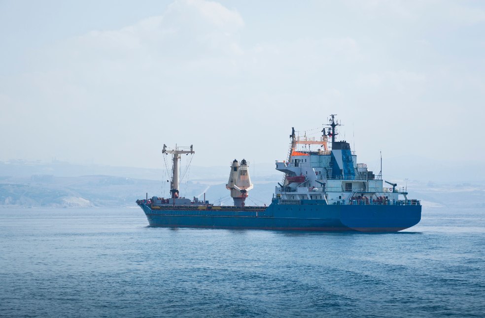 A large blue cargo container ship sails across the calm sea under a clear sky, symbolizing global trade