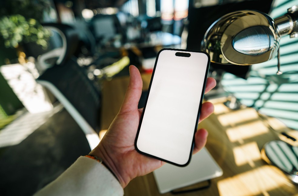 A hand holding a smartphone with a blank screen in a brightly lit modern office workspace.