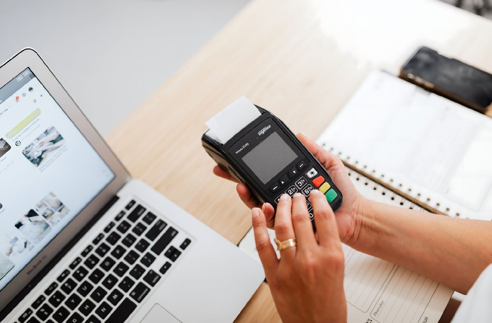 A person using a payment terminal with a laptop and a notebook on a work desk, illustrating cashless transactions.