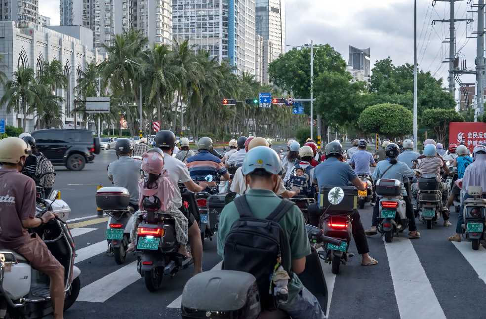 A crowded city intersection with motorcyclists stopped at a traffic light against a backdrop of modern buildings