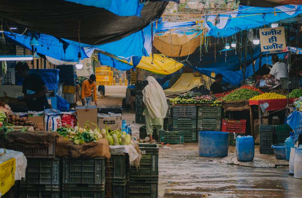 Colorful market scene with fresh produce and diverse shoppers under tarps.