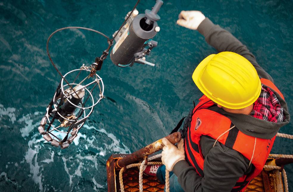 Worker in hard hat and life vest operates machinery on an offshore platform above ocean waters.