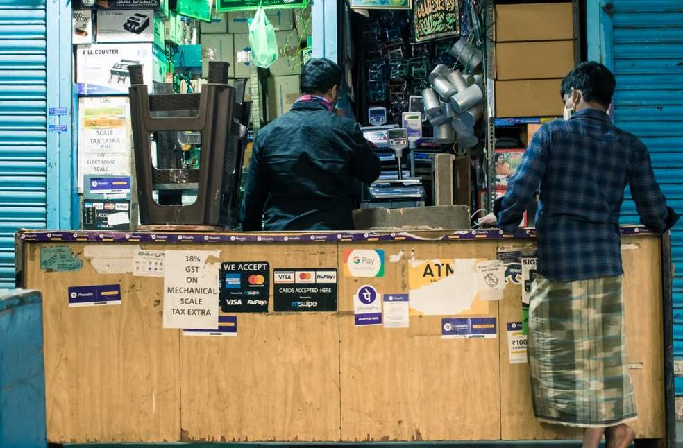 Two men at a shop counter in Bengaluru marketplace with payment signs and products displayed.