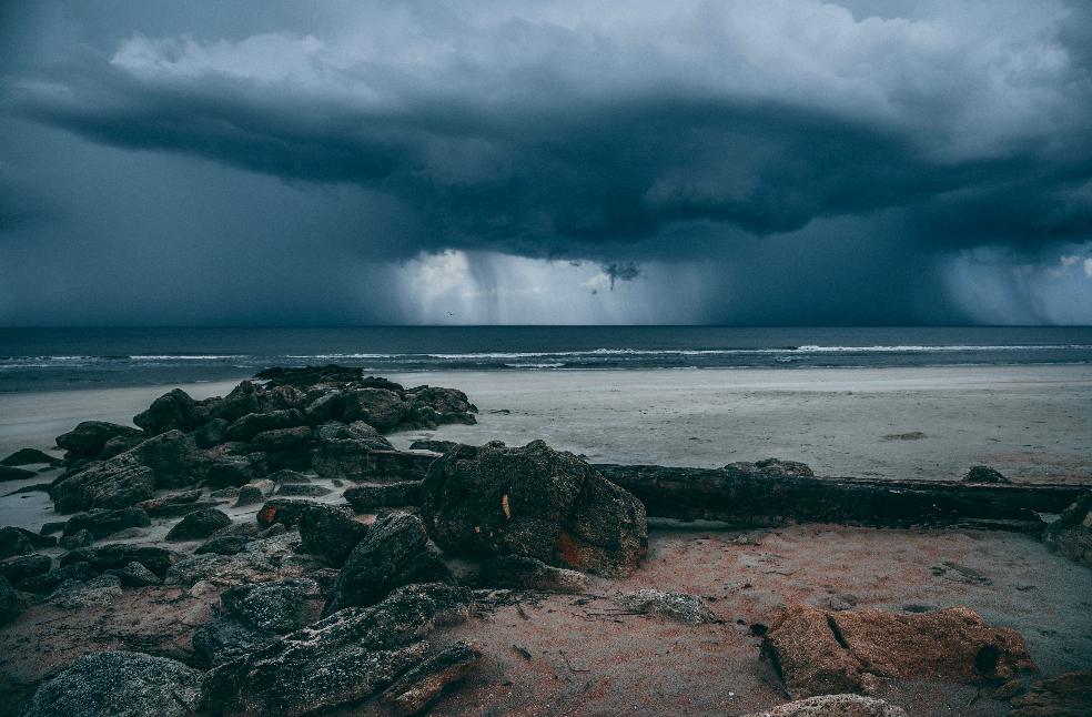 Moody seascape with storm clouds looming over rocky coastline and driftwood on sandy beach.