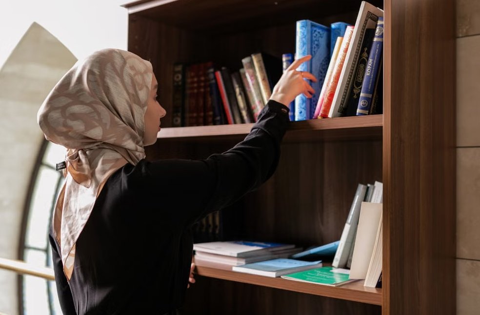 A woman in a headscarf reaches for a book on a wooden bookshelf in a serene indoor setting