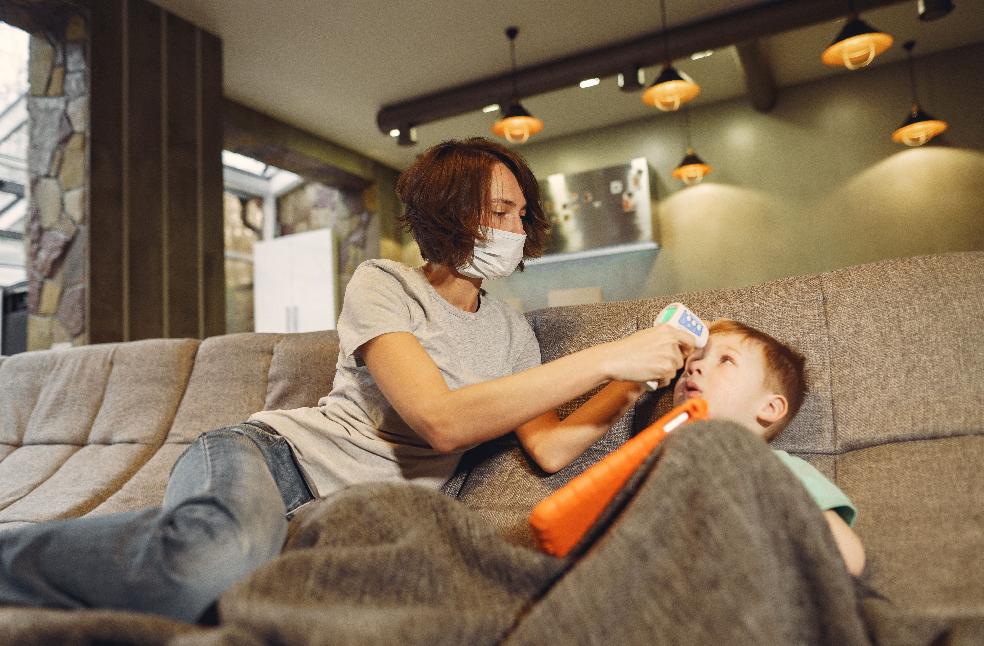 A mother checks her son's temperature at home using a digital thermometer, both practicing safety measures.