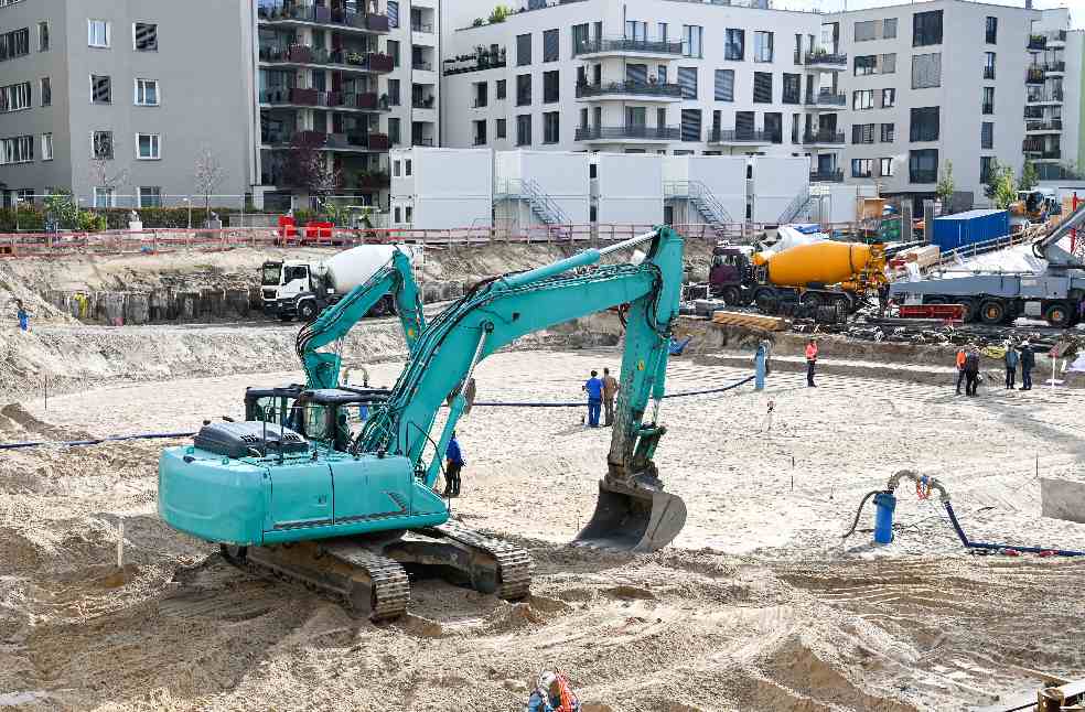 A busy construction site with machinery, workers, and residential buildings in the background.