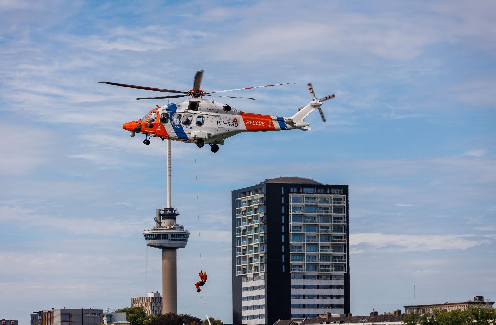 Coast guard helicopter conducting a rescue operation over Rotterdam with iconic Euromast tower in view.