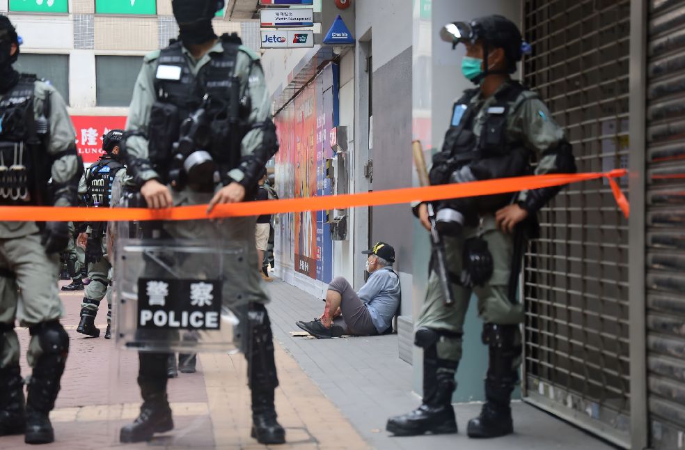 Urban environment with police presence and a civilian sitting against a wall, showing tension.