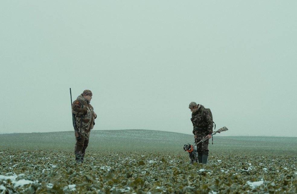 Two hunters with rifles and a dog in a snowy countryside field.