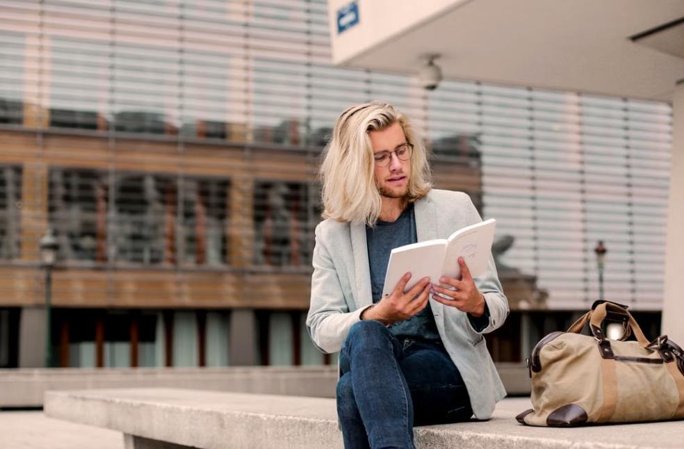 Man with long blond hair sitting on a bench outdoors, reading a book with a duffel bag beside him