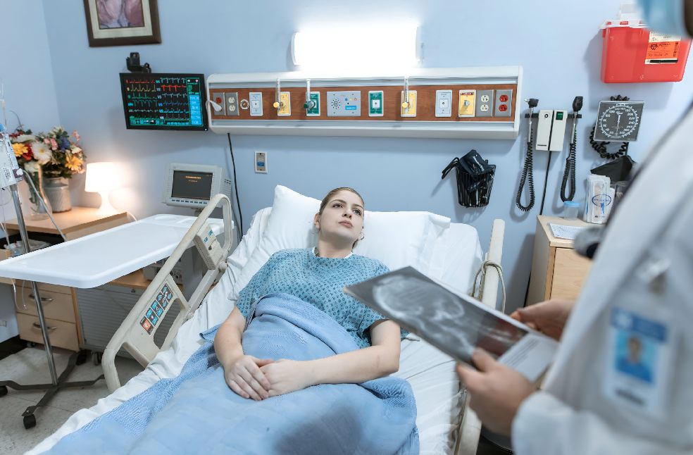 A female patient in a hospital gown receives medical attention from a healthcare professional holding an x-ray in a hospital ward.