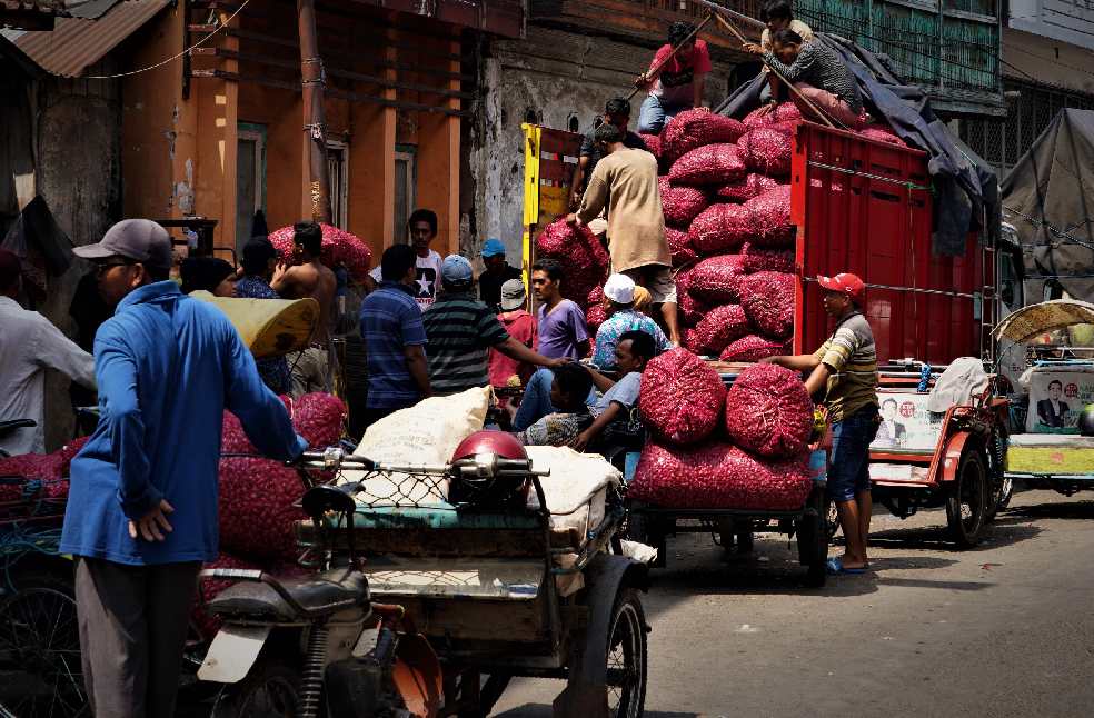 Men loading sacks onto a truck in a bustling market street filled with carts and people.