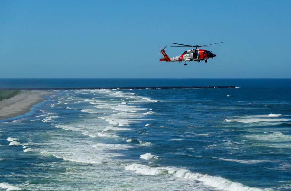 A Coast Guard helicopter flies above the ocean waves near the shore on a clear summer day.