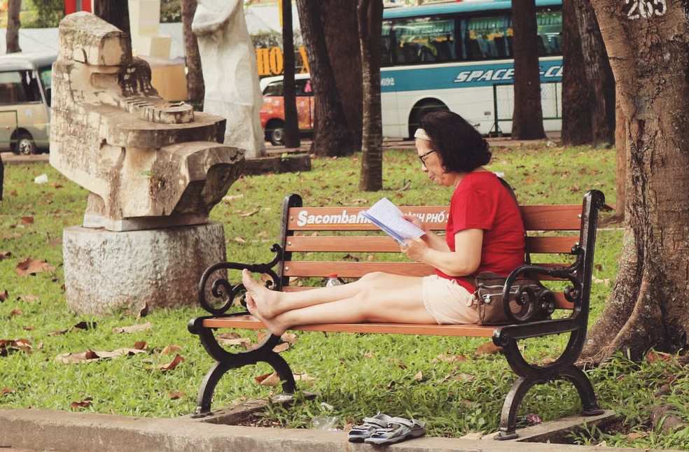 Woman relaxed on bench reading a book in an outdoor park setting with urban background
