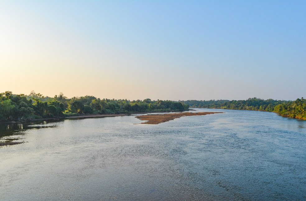 Calm river view with lush greenery and clear sky at sunset in Manipal, India.