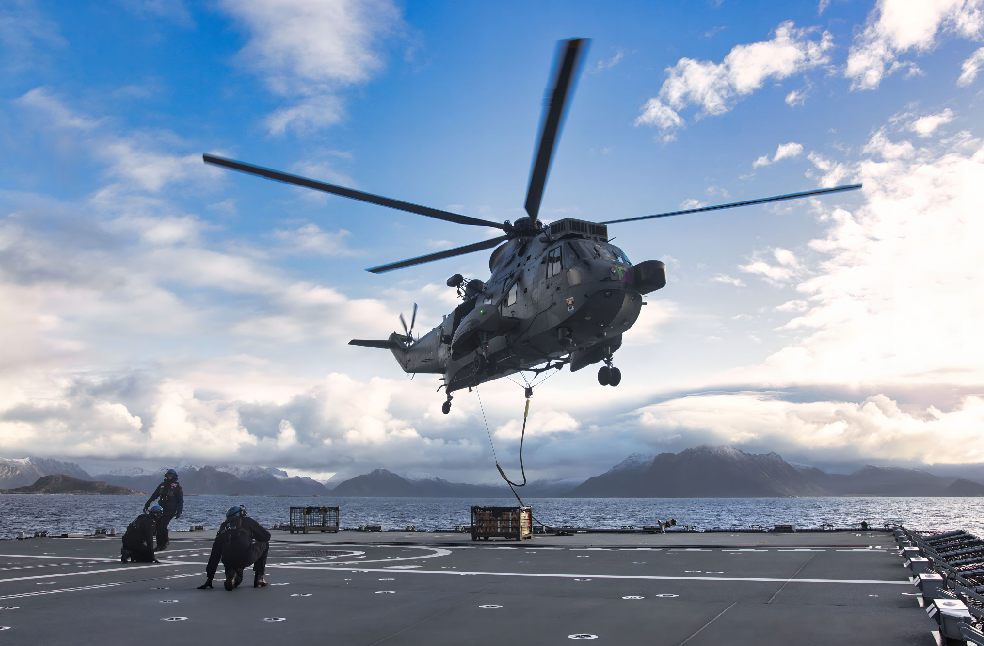 A military helicopter lands on a ship with mountains in the backdrop.
