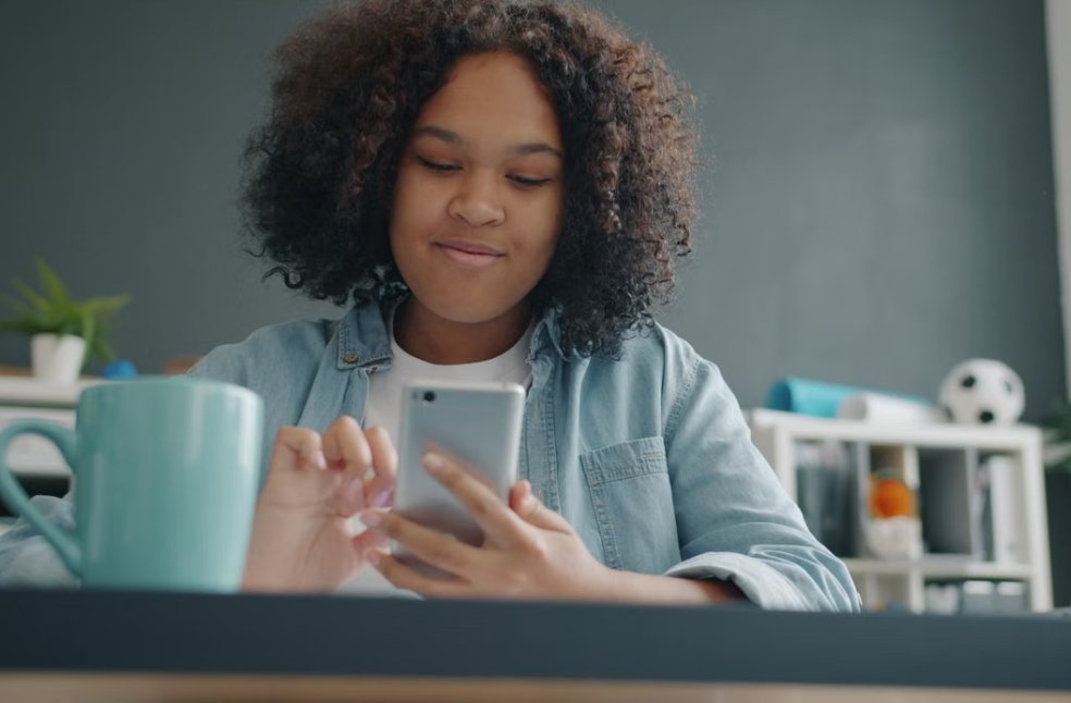 Teen girl enjoying time on smartphone in a cozy, modern room with a laptop and mug