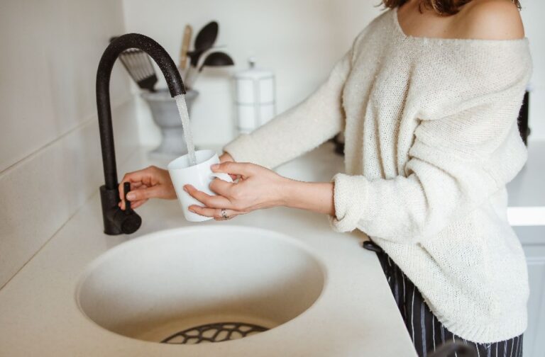US households face rising costs for drinking water service A woman filling a white cup with water from a black faucet in a modern kitchen.