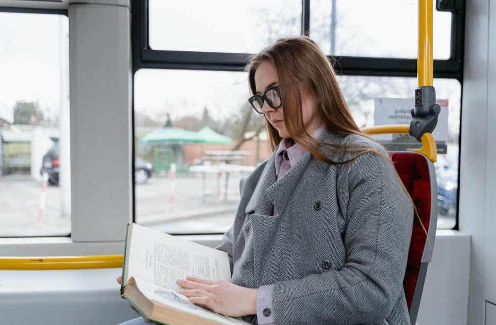 A young woman wearing eyeglasses reads a book while commuting on a bus